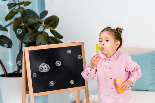 adorable child standing near blackboard and blowing soap bubbles  - Foto, Bild