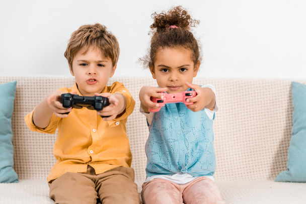 Adorables niños multiétnicos jugando videojuegos con joysticks y mirando a la cámara
 - Foto, imagen