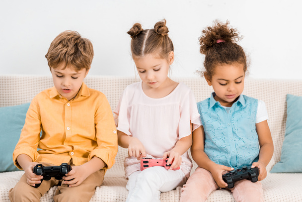 adorables niños multiétnicos sentados en el sofá y jugando con joysticks
 - Foto, Imagen