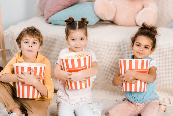 adorables niños pequeños sosteniendo cajas con palomitas de maíz y sonriendo a la cámara
 - Foto, Imagen
