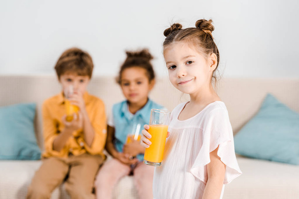 adorable niño sosteniendo un vaso de jugo y sonriendo a la cámara mientras pequeños amigos bebiendo jugo detrás
 - Foto, imagen