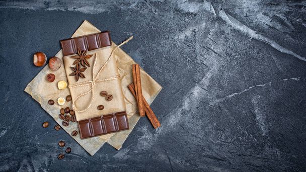 Top view of dark chocolate bar with coffee beans, whole hazelnuts and spices as cinnamon sticks and anise stars on black marble background with copy space - Photo, image