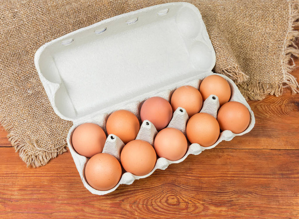Visão superior dos ovos de galinha marrom na caixa de ovos aberta para dez ovos feitos de polpa de papel reciclada em uma mesa rústica de madeira com pano de saco
 - Foto, Imagem