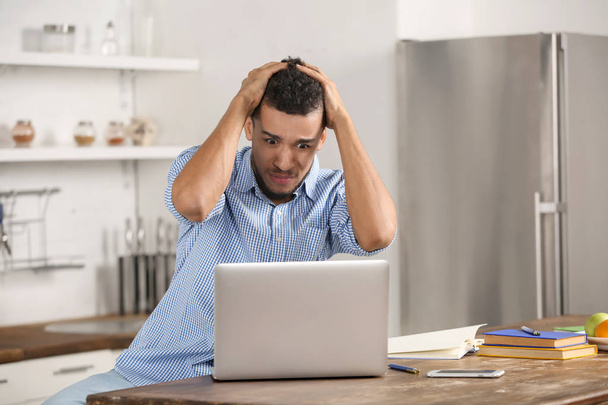 Stressed African-American man with laptop in kitchen - Фото, изображение