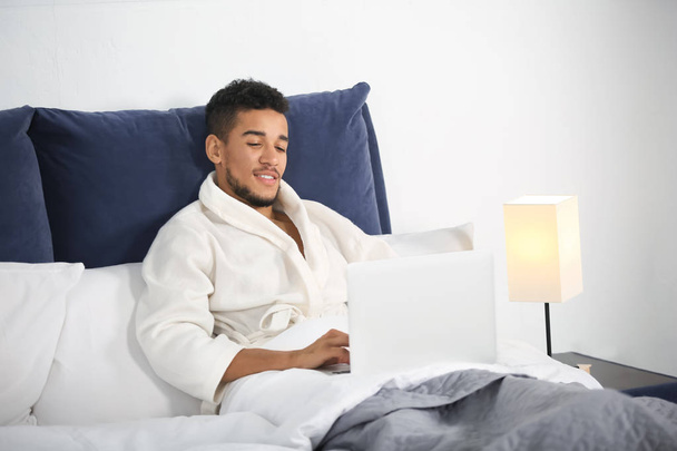 Young African-American man with laptop resting in bed - Fotografie, Obrázek