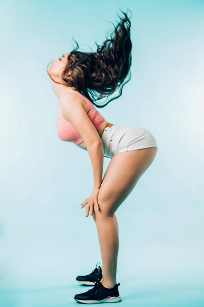 Attractive young brunette woman dancing on blue background. Sexy girl wears a pink top, white shorts, black crosses. Sports, healthy lifestyle, movement concepts - Φωτογραφία, εικόνα
