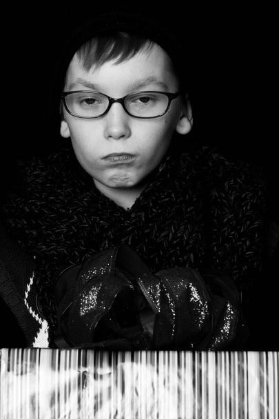 μικρό αγόρι σοβαρές ή χαριτωμένα nerd παιδί σε γυαλιά, καπέλο και μοντέρνο πλεκτό κασκόλ σε μαύρο φόντο κατέχει πολύχρωμο κουτί με κόκκινο bo - Φωτογραφία, εικόνα