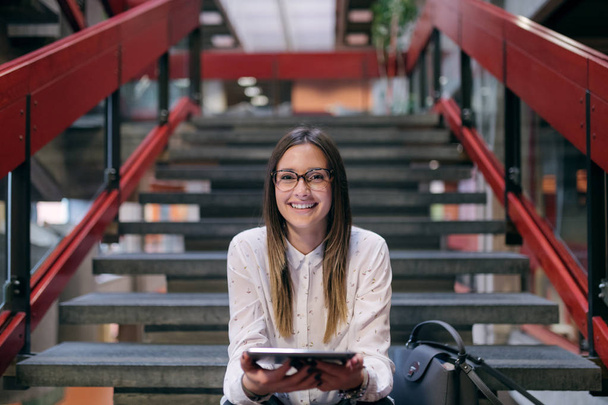 Молодая кавказская студентка с каштановыми волосами и очками, сидя на лестнице в здании колледжа, пользуется планшетом. Рядом с сумкой
. - Фото, изображение