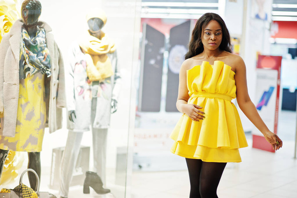 Stylish african american woman at yellow dreess posed against showcases with mannequins at mall. - Foto, afbeelding