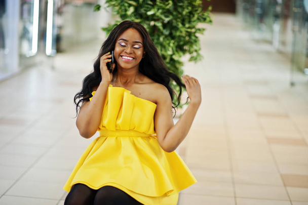 Stylish african american woman at yellow dreess posed at mall and sitting on bench inside, speaking at phone. - 写真・画像