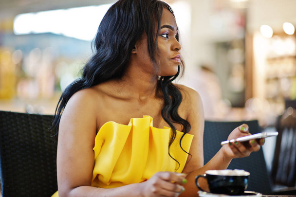 Stylish african american woman at yellow dreess posed at cafe with cup of cappuccino and mobile phone at hands. - Фото, изображение