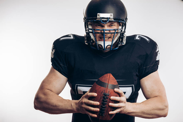 Muscular american football player in protective uniform and helmet holding ball - Фото, изображение