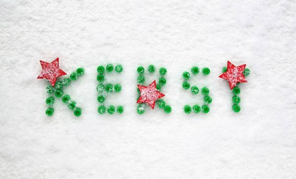 Weihnachtskarte in niederländischer Sprache. Schmuckschnur Perlen, grüne Buchstaben und rote Sterne. weißer schneebedeckter Hintergrund, Perspektive von oben und Textraum.  - Foto, Bild