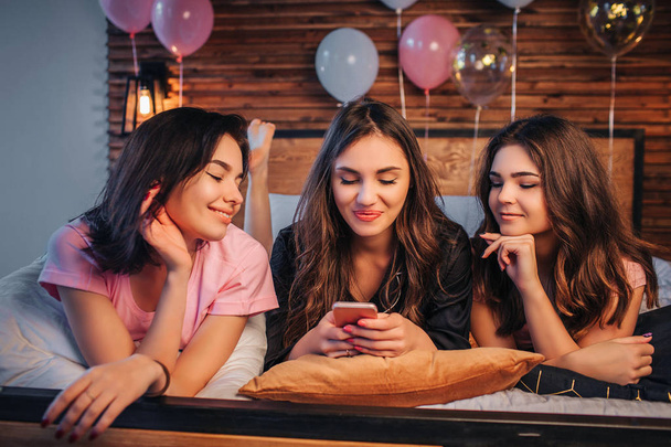 Three young women lying on bed in room. Girl in middle hold phone in hands. Her frieds look at it. They smile. Young women are positive and cheerful. - Foto, immagini