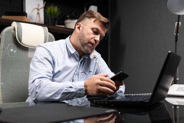 junger Mann sitzt am Computertisch und hält Festnetztelefon und Handy in der Hand - Foto, Bild