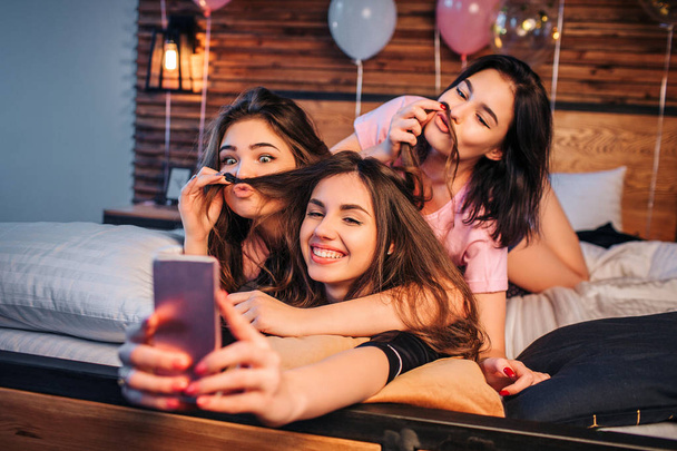 Three playful young women taking selfie on bed in room. Two models play with hair of third girl. They look on camera, pose and smile. Girls look happy. - Photo, Image
