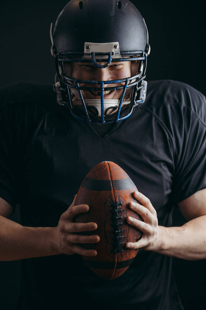American football player in black sportwear with a ball on black background - Foto, imagen