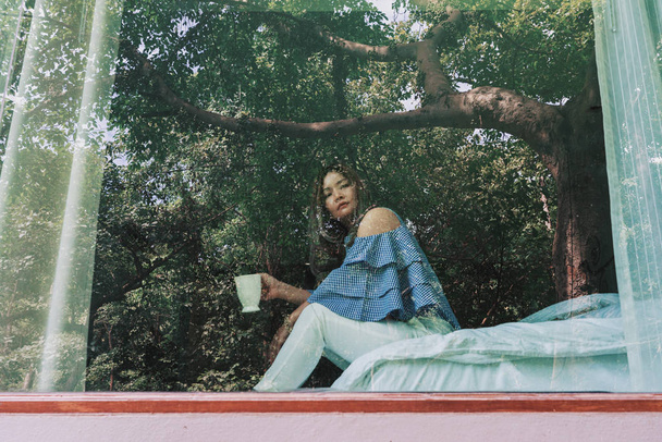 Pencere ağaçta yansıması ile beyaz Kupası onun elinde tutan yatağın üstüne oturan ufuk Asyalı kadın. - Fotoğraf, Görsel