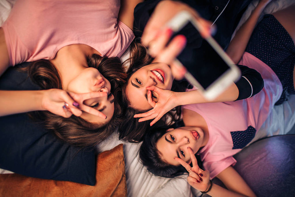 Three playful young women lying on bed. They pose on camera and make different poses. Model in middle hold white phone in hand. - Foto, afbeelding