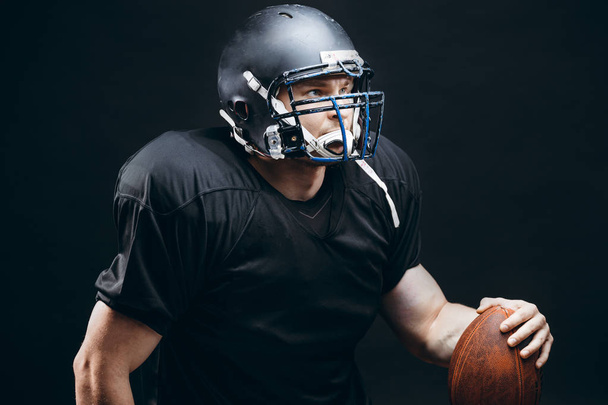 American football player in black sportwear with a ball on black background - Zdjęcie, obraz