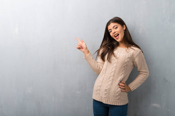 Menina adolescente com suéter em uma parede vintage apontando o dedo para o lado e apresentando um produto
 - Foto, Imagem