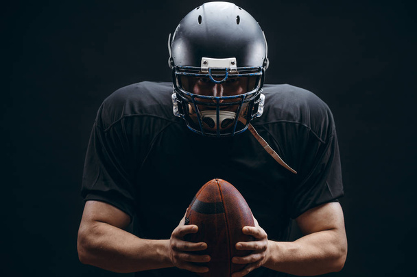 American football player in black sportwear with a ball on black background - Foto, Imagen