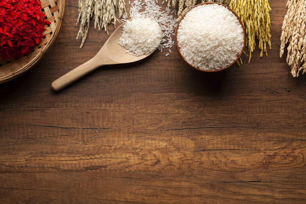 Vista superior del arroz jazmín en un tazón y cuchara de madera sobre una mesa de madera oscura con plantas de arroz, oreja de arroz con arroz jazmín en un tazón y flores rojas
. - Foto, imagen