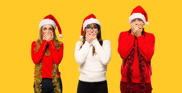 Μια ομάδα ανθρώπων ξανθιά γυναίκα που μεταμφιέζονται για τις διακοπές των Χριστουγέννων που καλύπτουν το στόμα για να πούμε κάτι ανάρμοστο. Δεν μπορεί να μιλήσει σε κίτρινο φόντο - Φωτογραφία, εικόνα