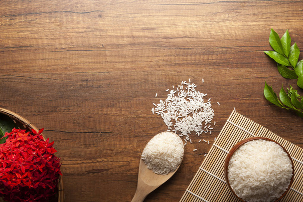 Vista dall'alto del riso al gelsomino in una ciotola e cucchiaio di legno su un tavolo di legno scuro con fiori rossi
. - Foto, immagini