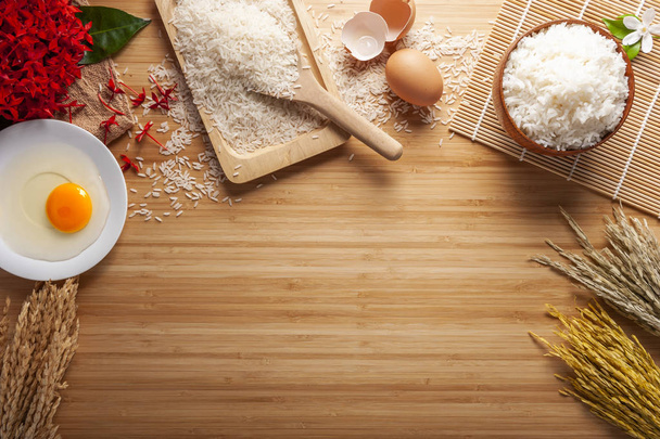 Vista superior de arroz de jasmim cozido em uma tigela em mesa de madeira escura com plantas de arroz e ovos, orelha de rices com arroz jasmim em uma tigela com flores vermelhas, dispersão de arroz no chão
. - Foto, Imagem