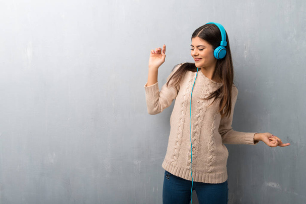 Menina adolescente com suéter em uma parede vintage ouvir música com fones de ouvido e dança
 - Foto, Imagem