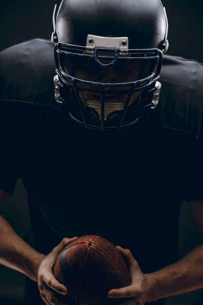 American football player with helmet and armour with a ball against black wall - Fotografie, Obrázek