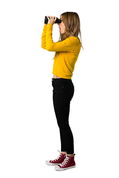 Un plano completo de una joven con suéter amarillo y mirando a lo lejos con prismáticos sobre fondo blanco aislado
 - Foto, Imagen