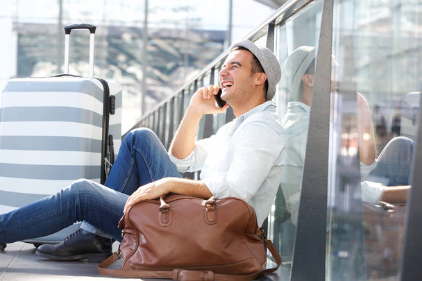 Side portrait of happy travel man sitting on floor with suitcase bags and talking on mobile phone at station - Фото, изображение