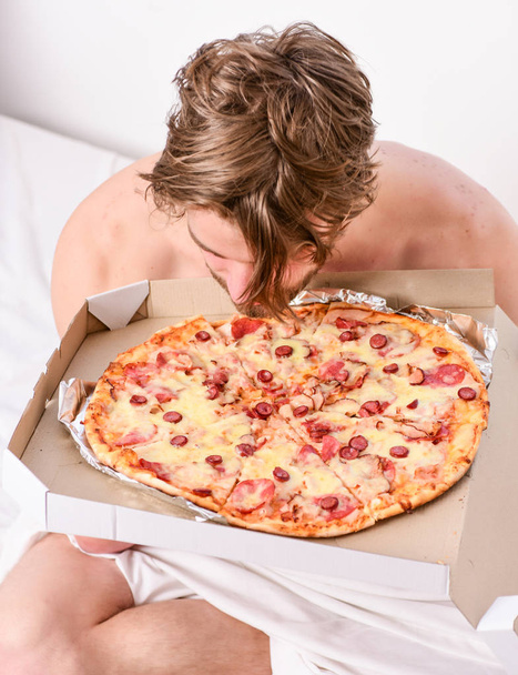 Man bearded handsome guy eating cheesy food for breakfast in bed. Shirtless handsome young man with pizza on bed. Student is at home on the bed in a bright apartment eating a tasty pizza - Fotografie, Obrázek