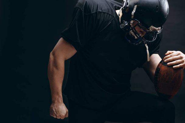 Giocatore di football americano con casco e armatura con palla contro muro nero - Foto, immagini