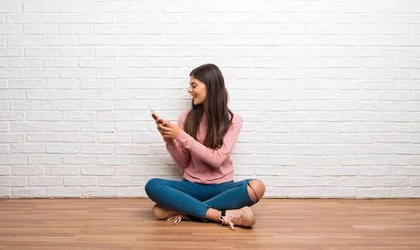 Девушка-подросток сидит на полу в комнате и отправляет сообщение с мобильного телефона.
 - Фото, изображение
