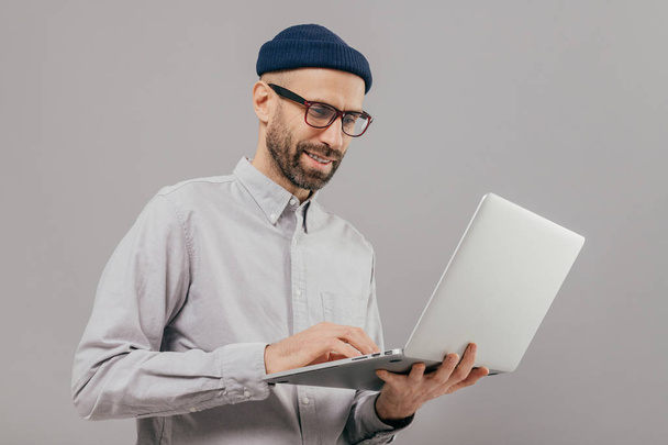 Снимок довольного мужчины с густой бородой и усами, держит ноутбук, переводит деньги, использует онлайн-банкинг, отправляет файлы, подключенные к беспроводному интернету, модели на сером фоне
 - Фото, изображение