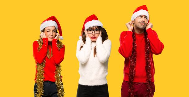 Μια ομάδα ανθρώπων ξανθιά γυναίκα που μεταμφιέζονται για τις διακοπές των Χριστουγέννων που καλύπτουν τα αυτιά με τα χέρια. Απογοητευμένοι έκφραση σε κίτρινο φόντο - Φωτογραφία, εικόνα