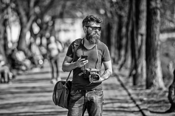 Bearded man carries bag on shoulder and holds camera. Photographer concept. Man with long stylish beard using smartphone while walking. Man with beard on confident face, urban background, defocused - Φωτογραφία, εικόνα