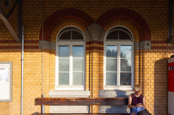 young lady sitting on bench at trainstation before brick wall in bavaria south germany hot summer day - Fotó, kép