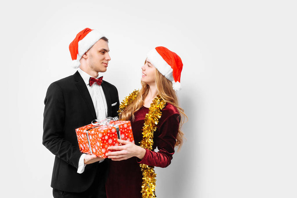 A happy couple of lovers, in Santa Claus hats, looking at each other, with Christmas gifts in their hands. On a white background. - Zdjęcie, obraz