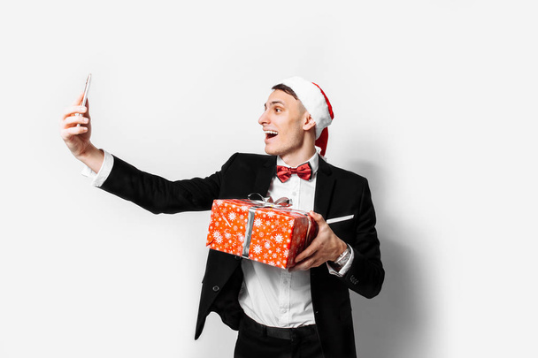 Shocked guy in a suit and a santa hat, in his hands - a Christmas gift, he takes pictures of himself on a smartphone. On a white background. - Foto, Imagen