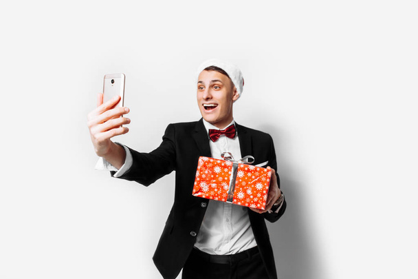 Shocked guy in a suit and a santa hat, in his hands - a Christmas gift, he takes pictures of himself on a smartphone. On a white background. - Fotografie, Obrázek