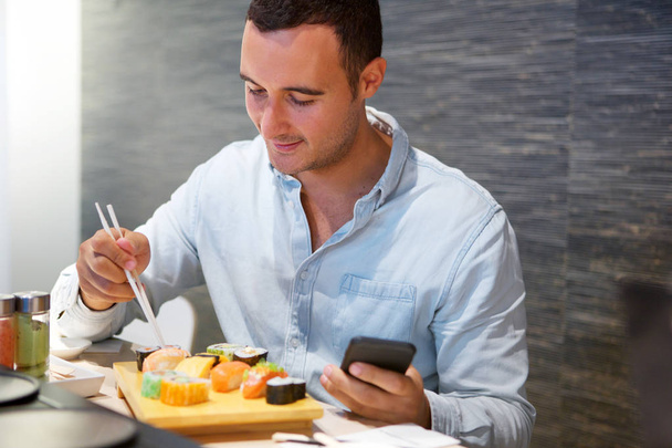 Portrait of man sitting at sushi restaurant with cellphone - 写真・画像