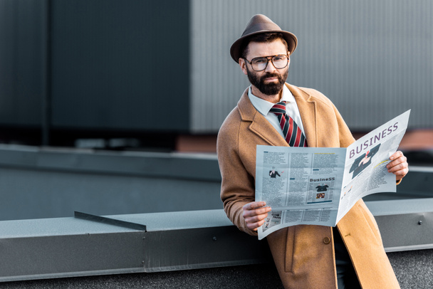 handsome adult man in formalwear holding business newspaper and looking at camera - Zdjęcie, obraz