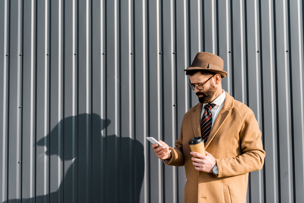 businessman in coat and glasses using smartphone and holding coffee to go near wall with shadow - Fotografie, Obrázek