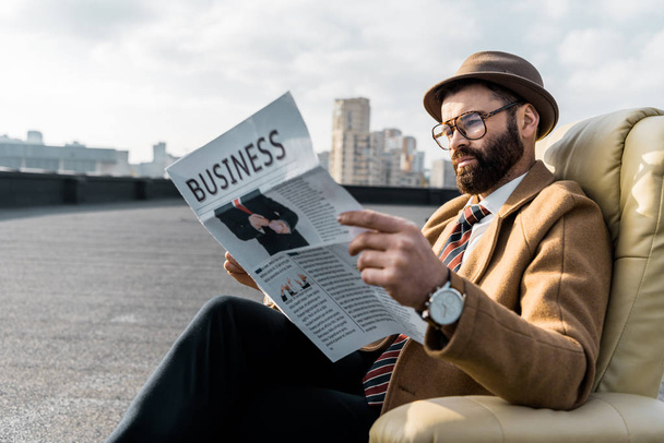 bearded man in glasses reading business newspaper in armchair on roof - Фото, зображення