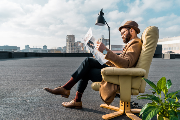 γενειοφόρος επιχειρηματίας συνεδρίαση στην πολυθρόνα και διαβάζει εφημερίδα στη στέγη  - Φωτογραφία, εικόνα