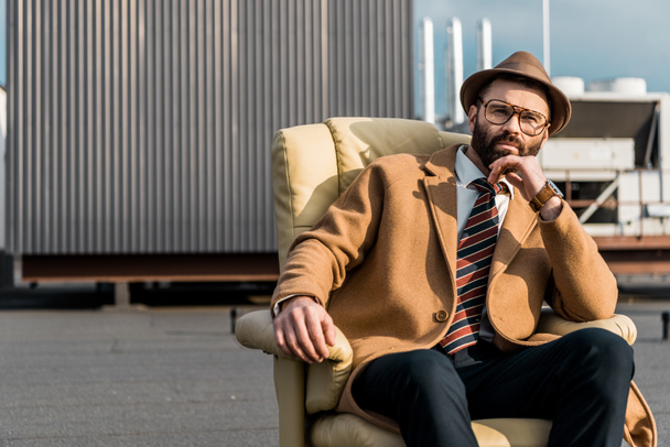 adult businessman in hat and glasses sitting in armchair on roof  - Valokuva, kuva
